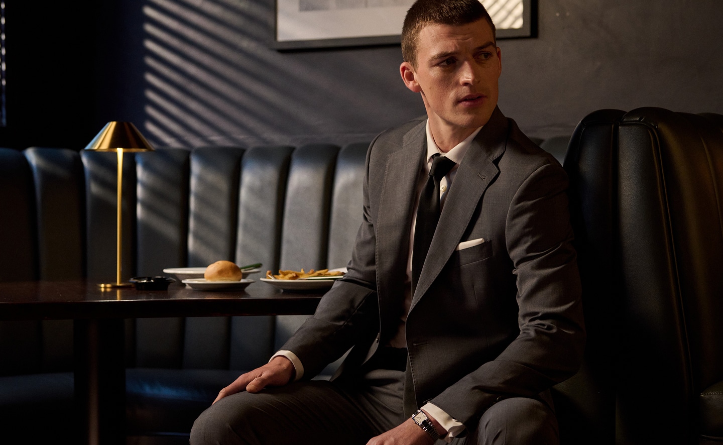 Man in a charcoal suit seated in a restaurant booth. Online only suits up to 50 percent off.