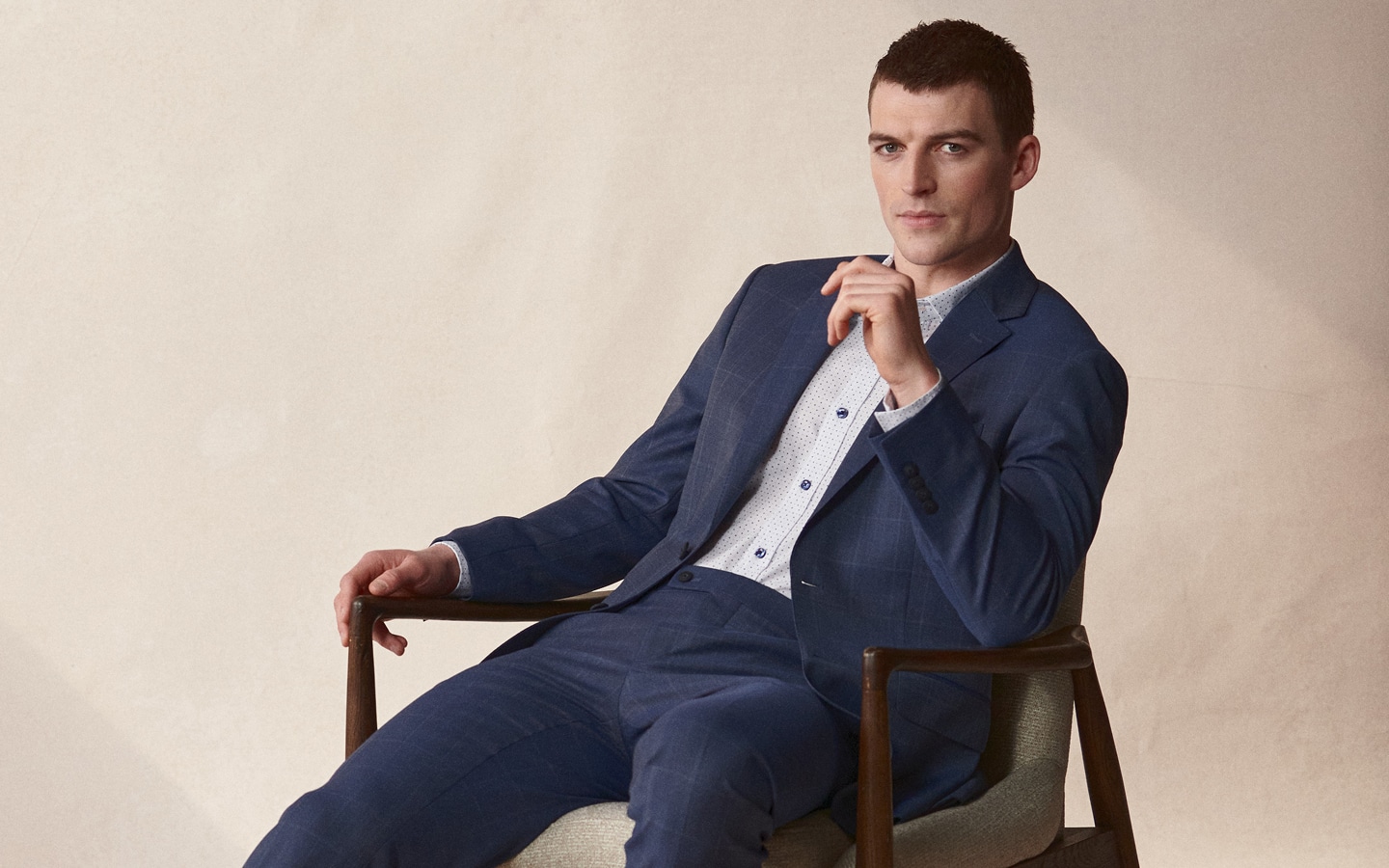 Suits up to 50% off. Man in tailored navy patterned suit sitting in a mid-century modern chair.
