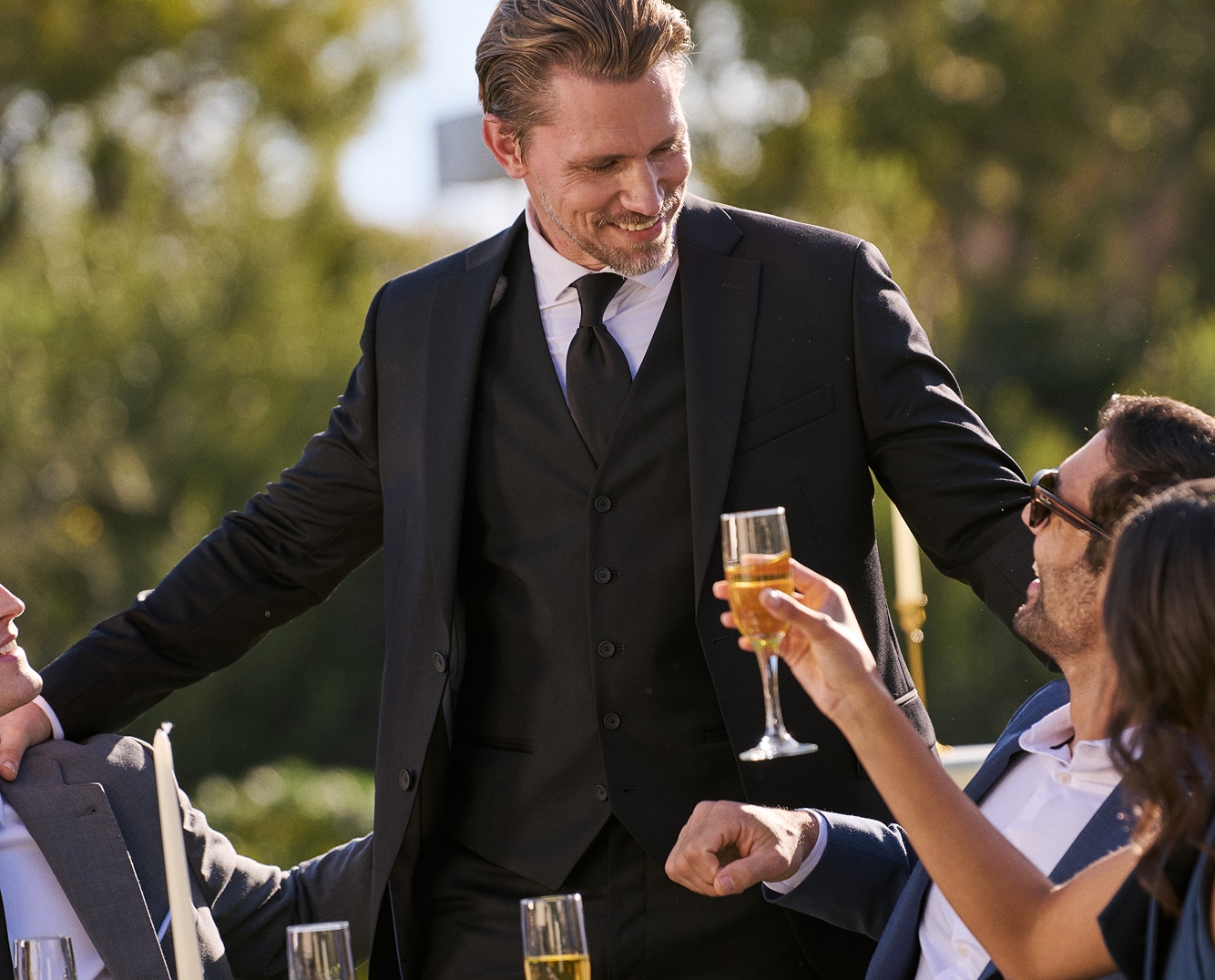 Man in a black three-piece suit at an outdoor wedding reception.