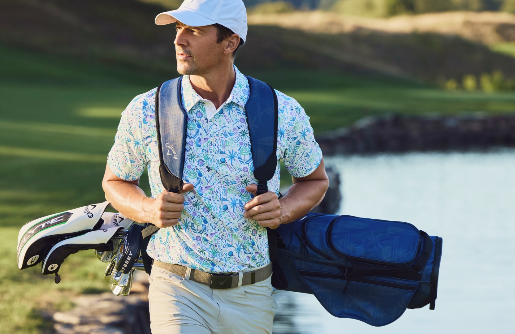 Man on a golf course carrying a golf bag, wearing a patterned golf polo and cap