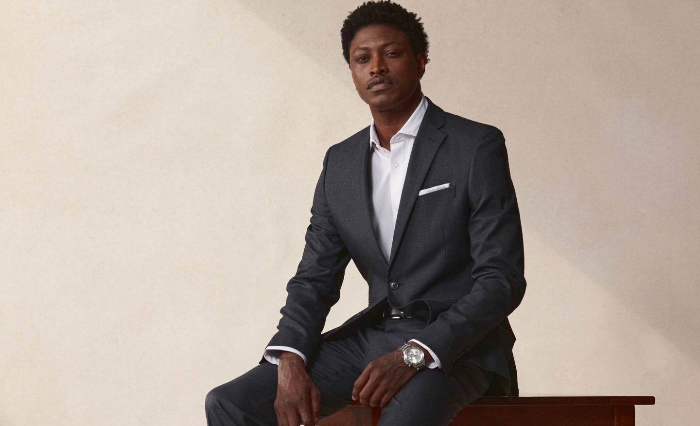 Suits 30% off. Man in tailored navy patterned suit sitting in a mid-century modern chair.