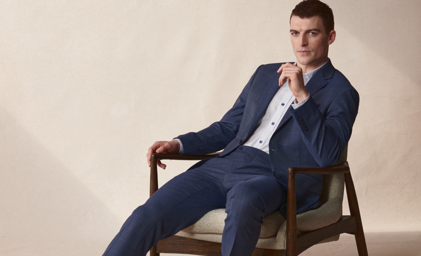 Suits 30% off. Man in tailored navy patterned suit sitting in a mid-century modern chair.